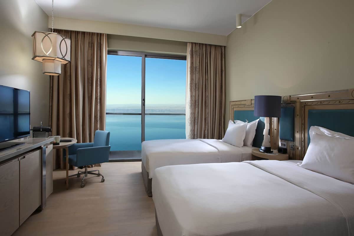 A hotel room with two beds, a desk with a TV, a chair, and a view of the sea.