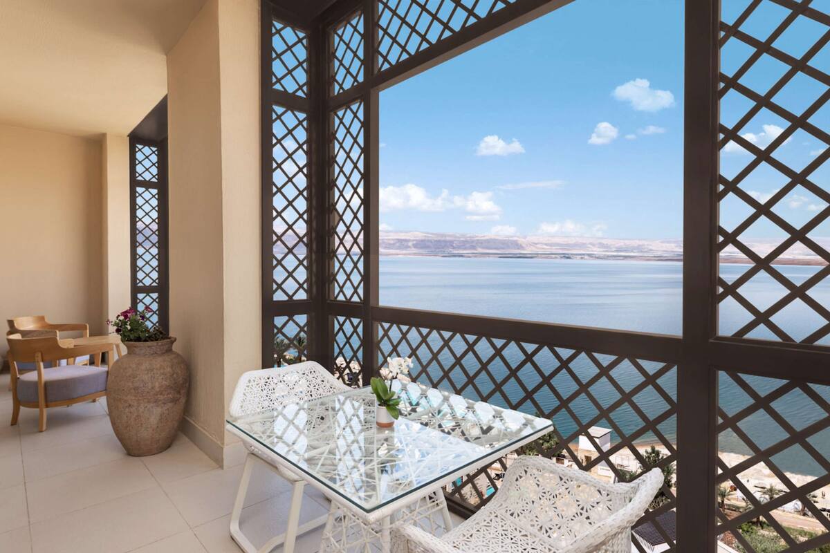 A balcony with a glass table and white wicker chairs, overlooking a body of water and distant mountains.