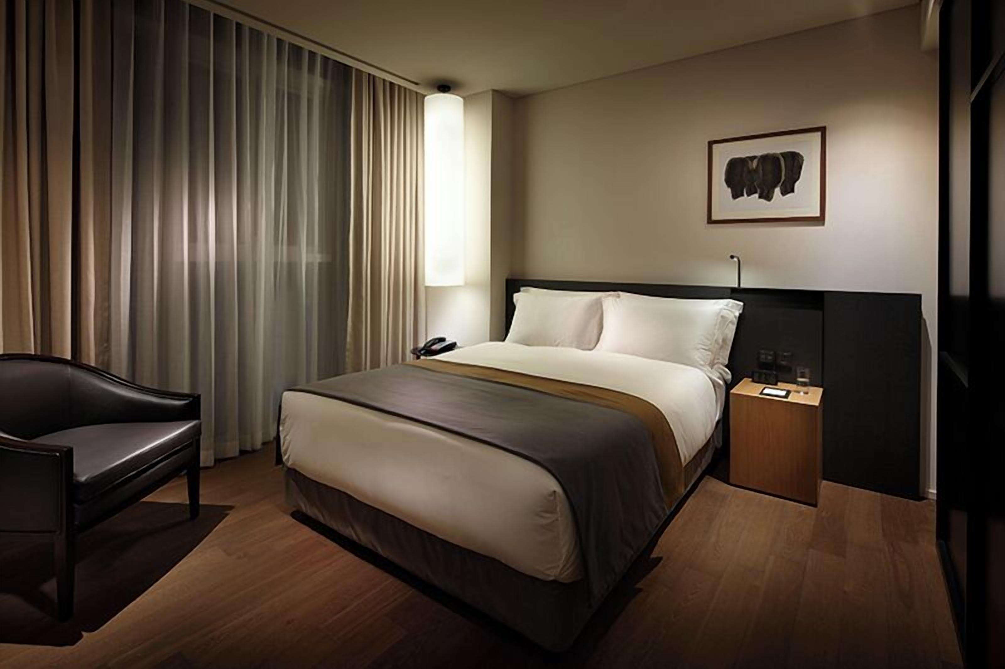 A hotel room with a bed, a chair, a bedside table with a lamp, a phone, and a framed picture on the wall.
