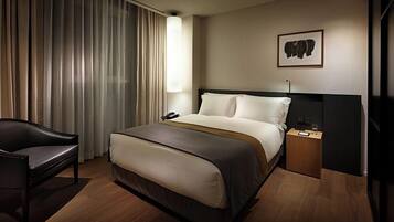 A hotel room with a bed, a chair, a bedside table with a lamp, a phone, and a framed picture on the wall.