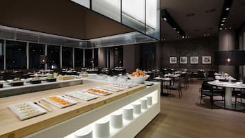 A modern restaurant with a long counter displaying various dishes, multiple dining tables, and a large window overlooking the cityscape.