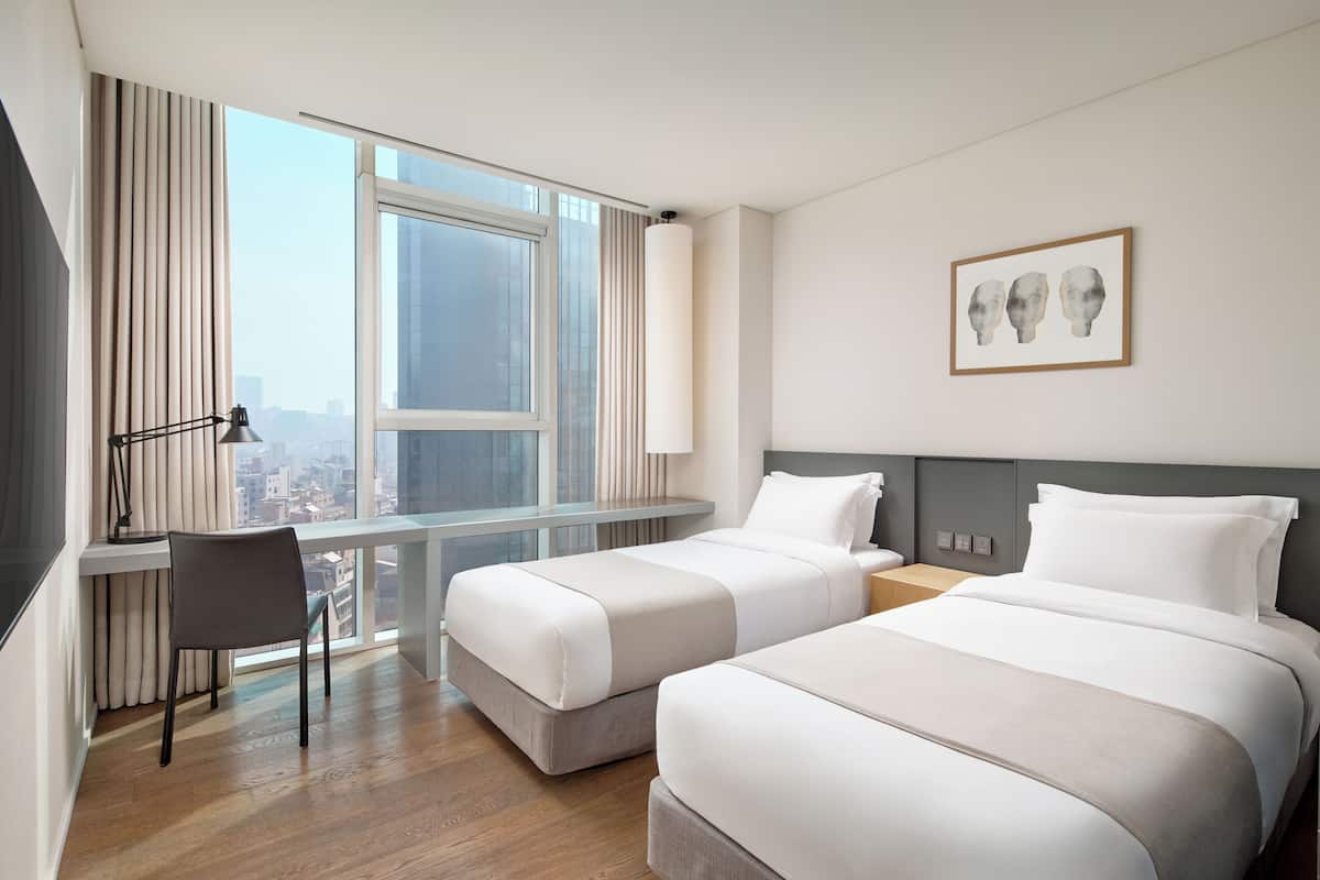 Habitación de hotel con dos camas, un escritorio, una silla, una lámpara y ventana con vista a la ciudad.