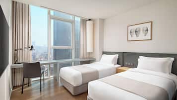 Quarto de hotel com duas camas, uma escrivaninha, uma cadeira, um abajur e uma janela com vista para a cidade.