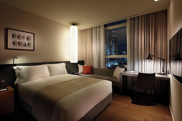 Habitación de hotel con una cama grande, un escritorio, una silla, ventana con cortinas y vistas nocturnas de la ciudad.