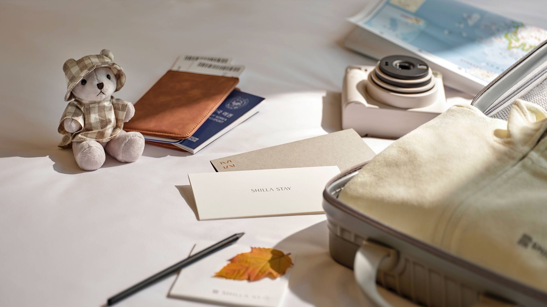 A travel setup with a teddy bear, a wallet, a passport, a camera, a notebook, a pen, a leaf, and a suitcase on a bed.
