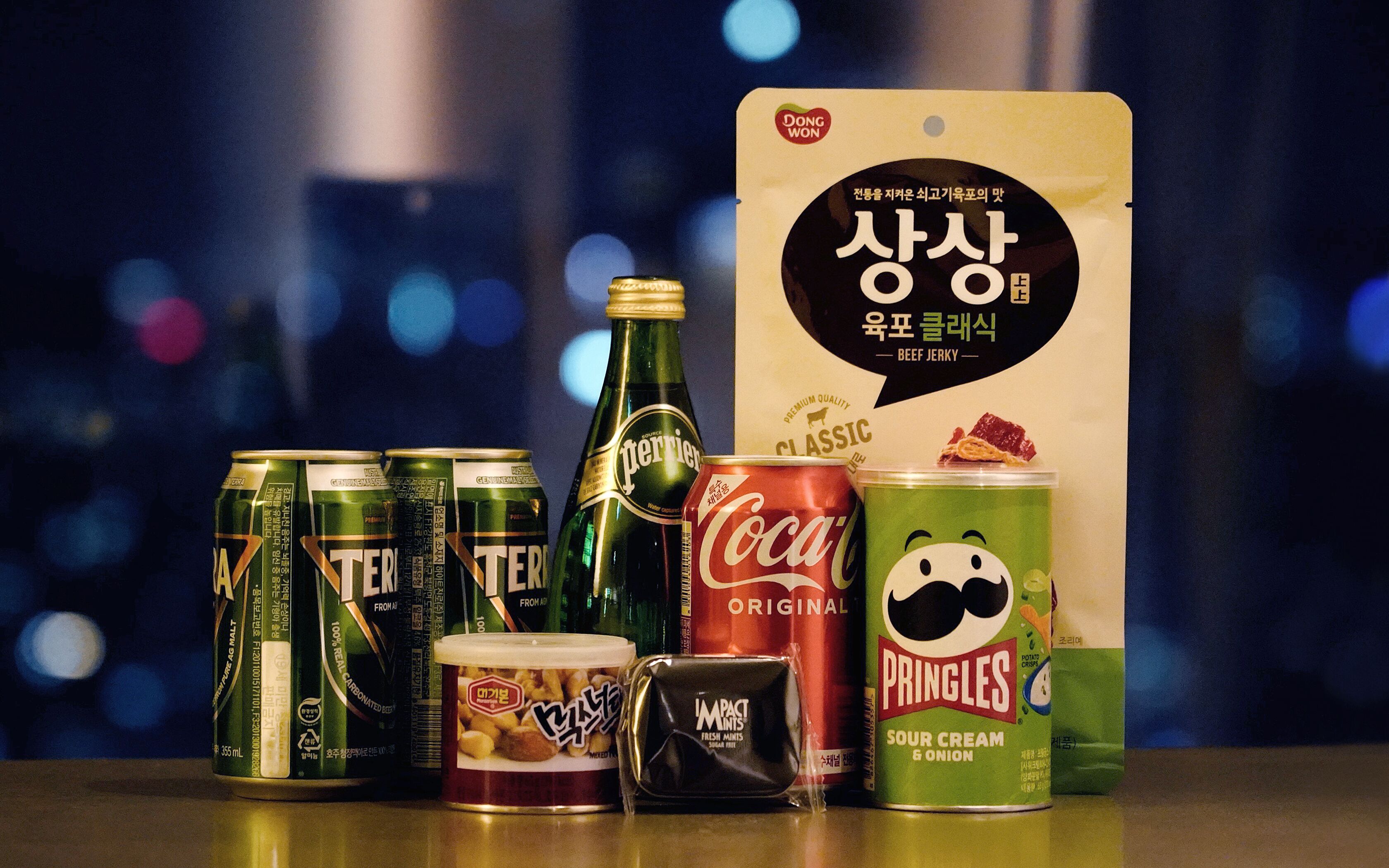 Variedad de alimentos y bebidas, incluyendo latas de cerveza, una botella de agua Perrier, una lata de Coca-Cola, una lata de Pringles y un paquete de carne seca.