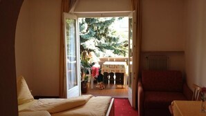 A room with a bed, a red sofa, a table with a vase, and a view of a dining area with a table and chairs. - Pension Schweitzer (Silandro)
