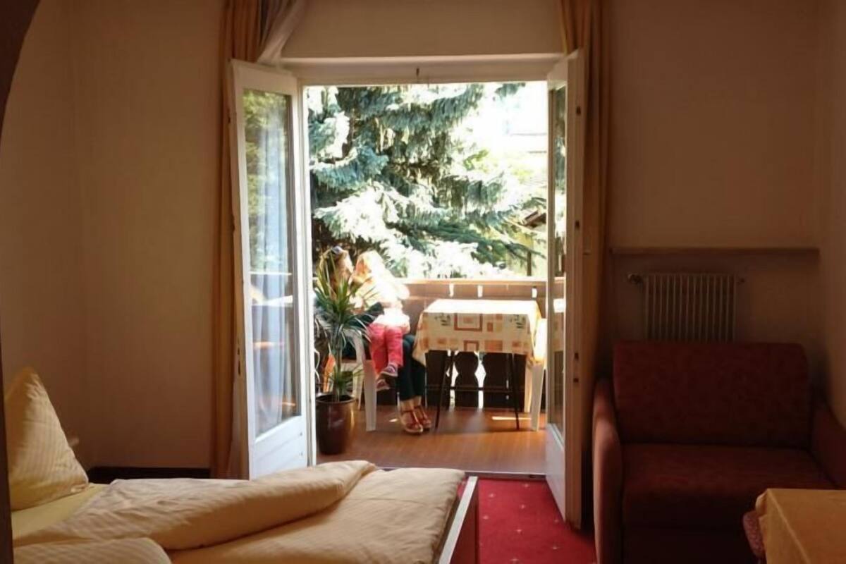 A room with a bed, a red sofa, a table with a vase, and a view of a dining area with a table and chairs.