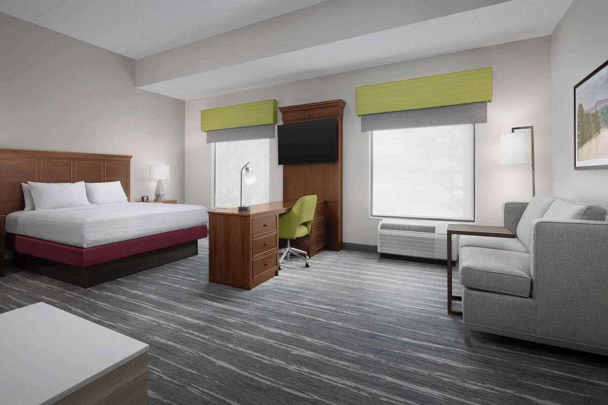 A hotel room with a bed, desk, chair, sofa, and large windows.