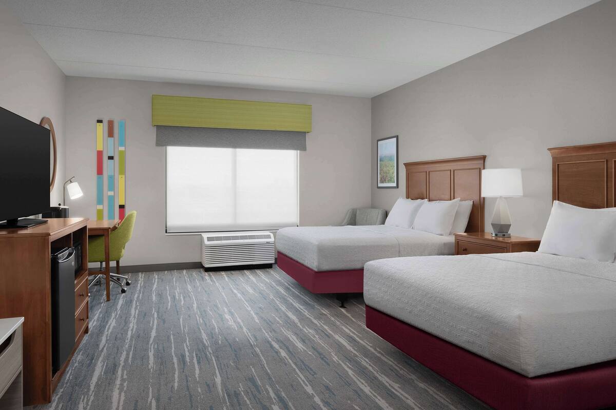 A hotel room with two beds, a desk with a chair, a television, and a window with blinds.
