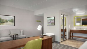 Une chambre d’hôtel moderne avec un bureau en bois, une chaise verte, un canapé, une petite table, une lampe de bureau, un lit, une salle de bain avec un miroir et deux tableaux encadrés accrochés aux murs.