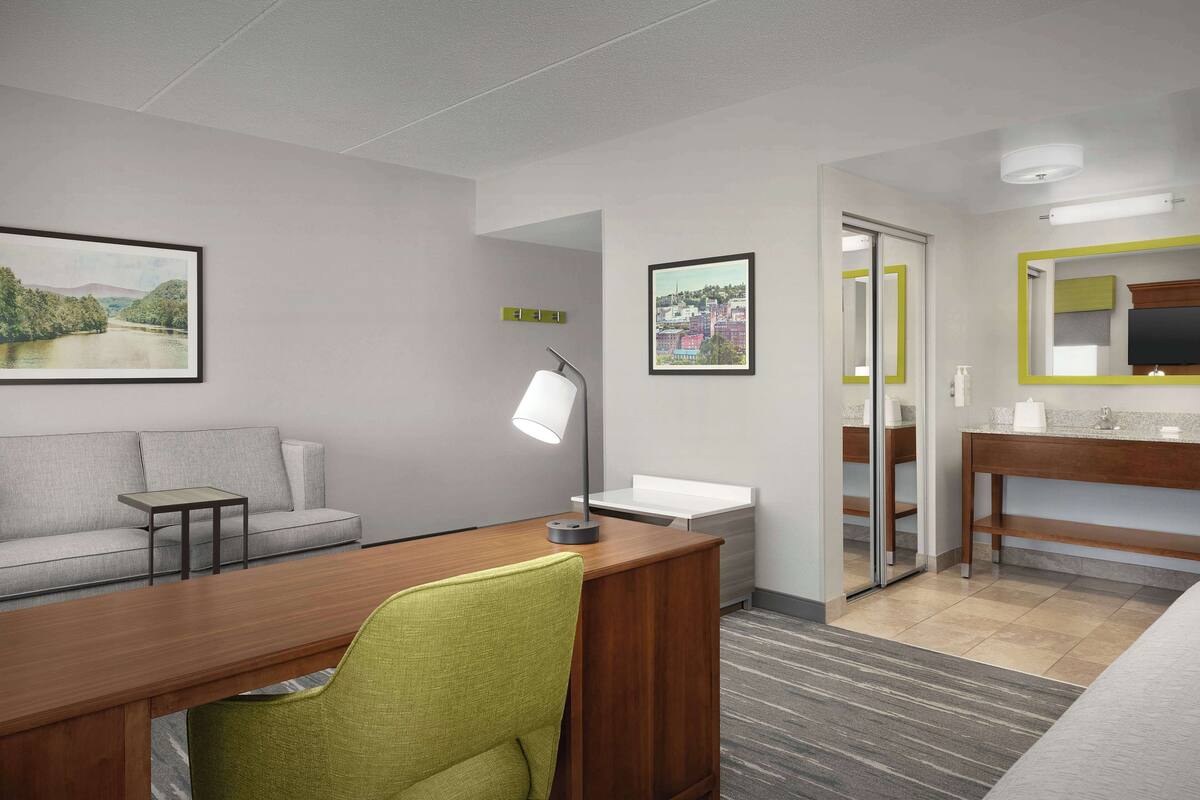 A modern hotel room with a wooden desk, a green chair, a sofa, a small table, a desk lamp, a bed, a bathroom with a mirror, and two framed pictures on the walls.