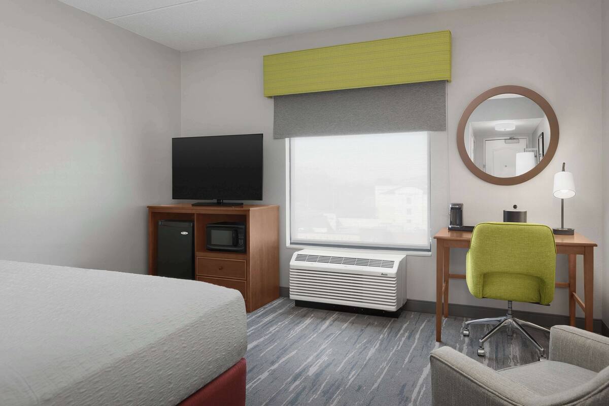 A hotel room with a bed, a desk with a chair, a television, an air conditioner, and a window with a view.