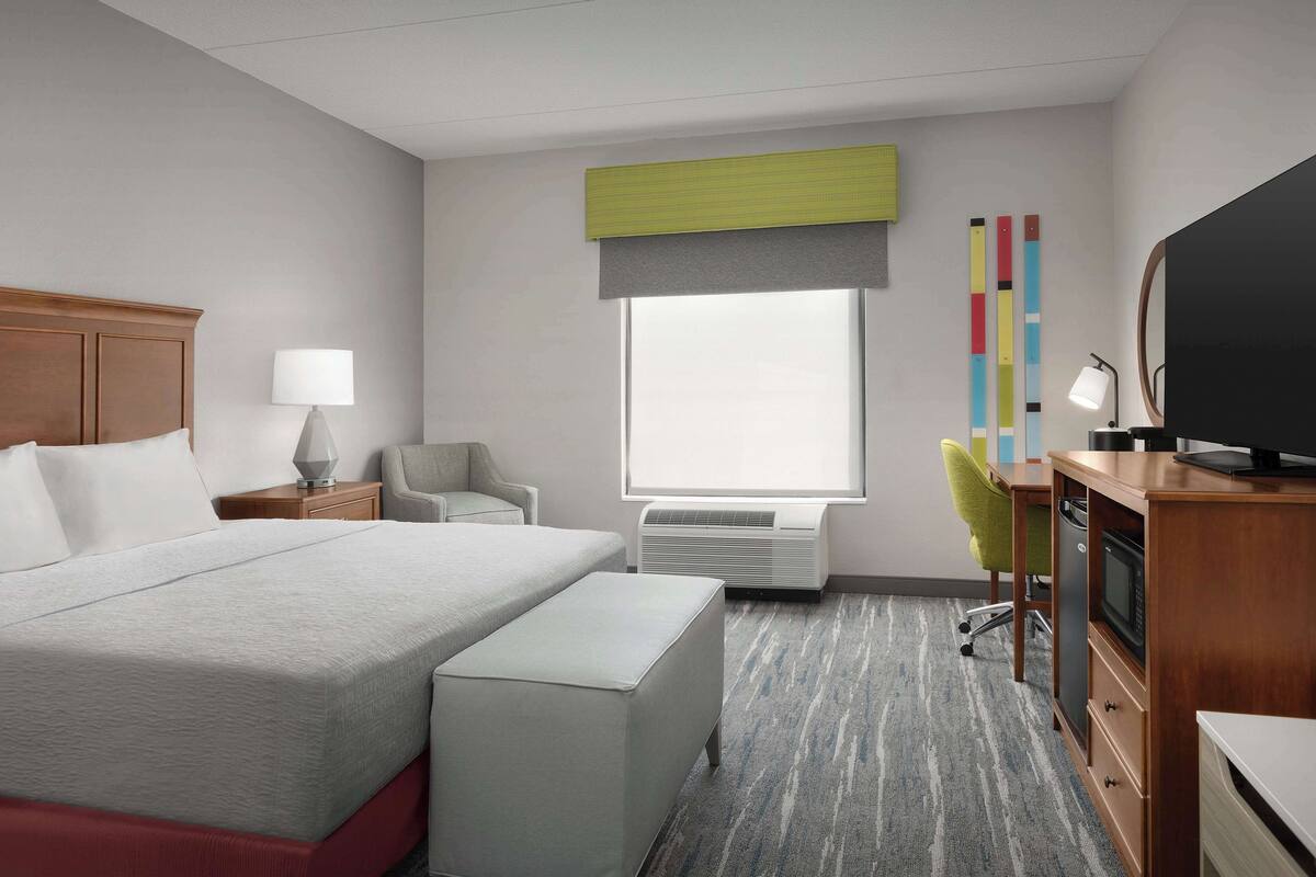 A hotel room with a large bed, a desk with a television, a chair, a lamp, and a window with a view of colorful vertical blinds.