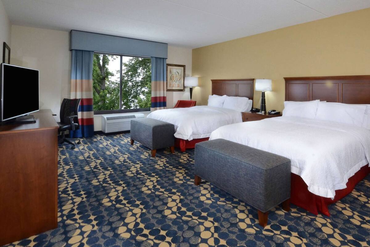 A hotel room with two beds, a TV, a desk, and a window with curtains.