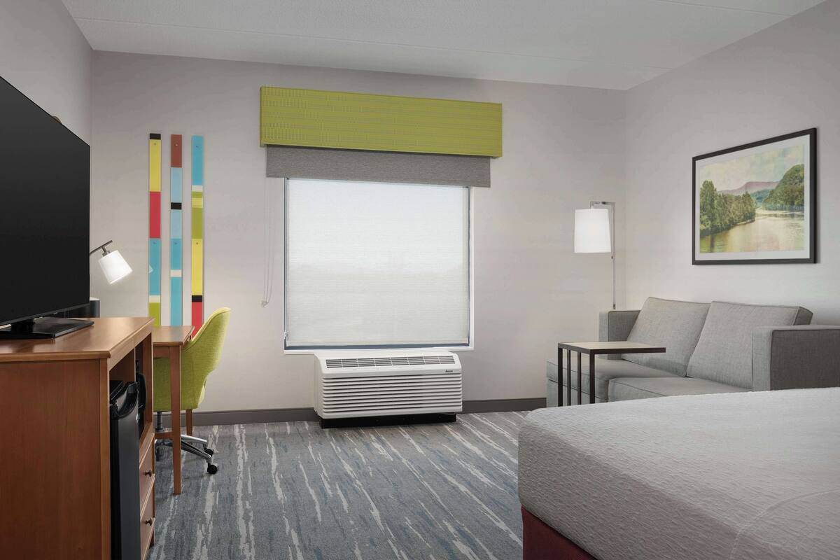 A hotel room with a bed, a desk with a chair, a TV, a window with blinds, a painting on the wall, and a heater.