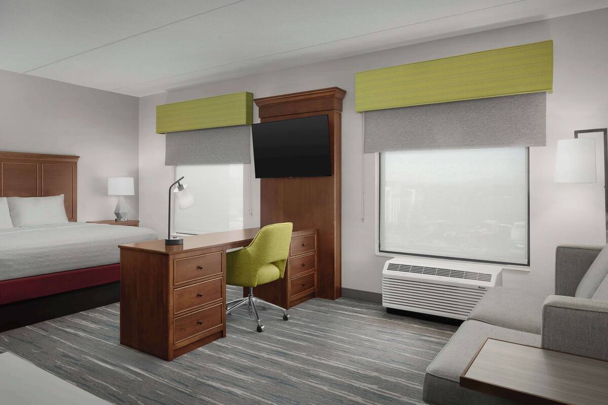 A hotel room with a bed, a desk with a chair, a TV, a window with blinds, and an air conditioning unit.