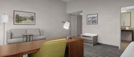 Habitación de hotel con escritorio de madera, una silla verde, un sofá gris, una mesita, una mesita de noche, una lámpara y dos cuadros en las paredes.