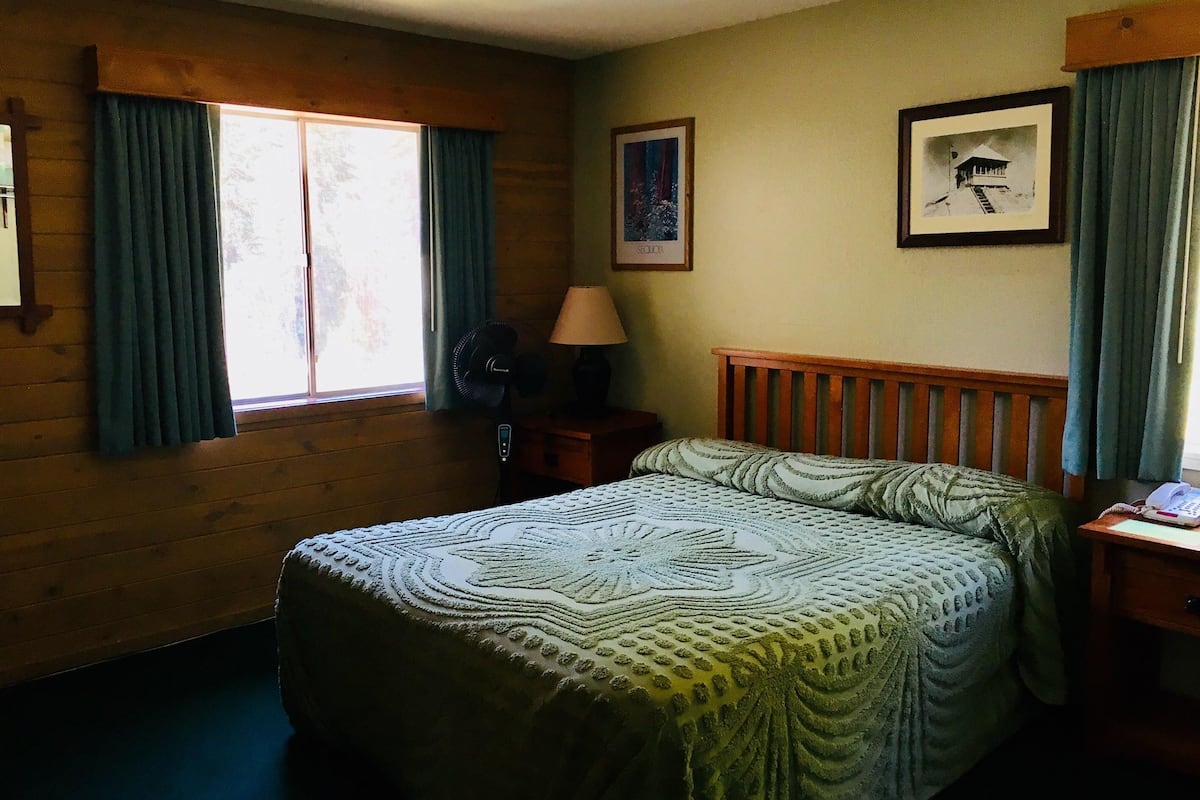 Una habitación con una cama de madera, un ventilador de pie, una lámpara y un cuadro en la pared.