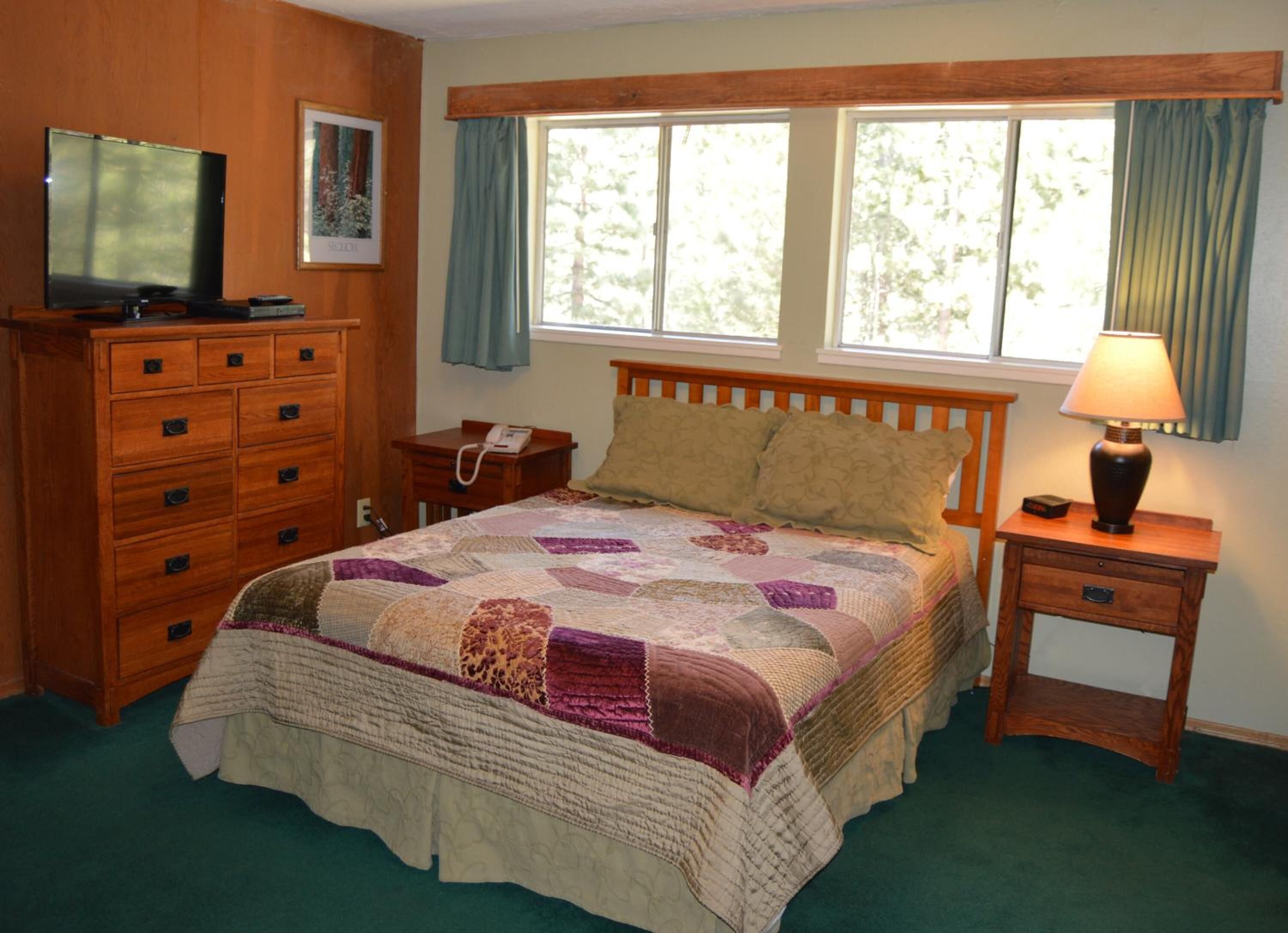 A bedroom with a bed, wooden dresser, TV, bedside lamp, and a window with curtains.