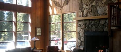 A cozy lodge interior with large windows, a stone fireplace, wooden furniture, and a ceiling fan.