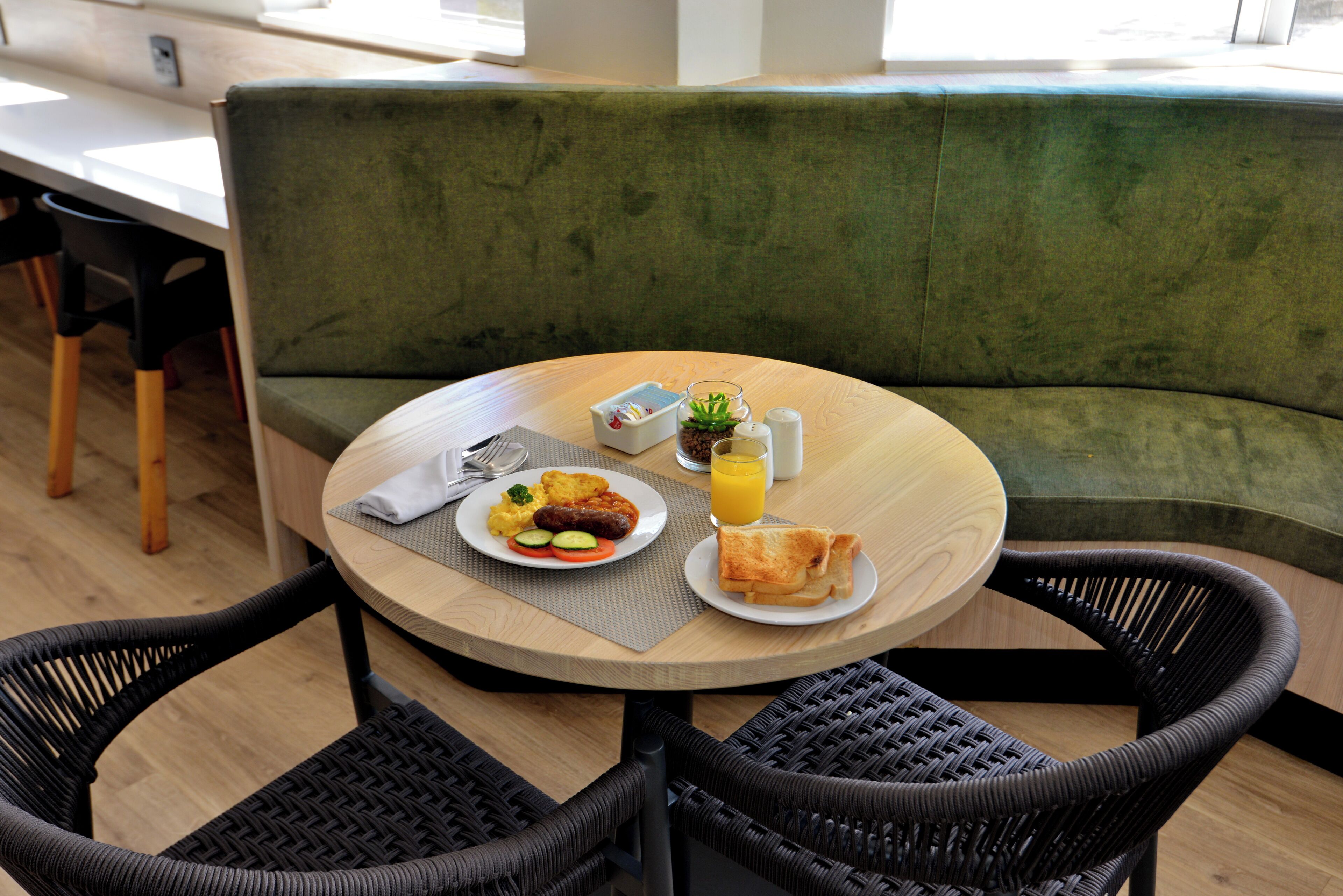 A round wooden table with breakfast items including eggs, sausages, and toast, set on a green upholstered bench.