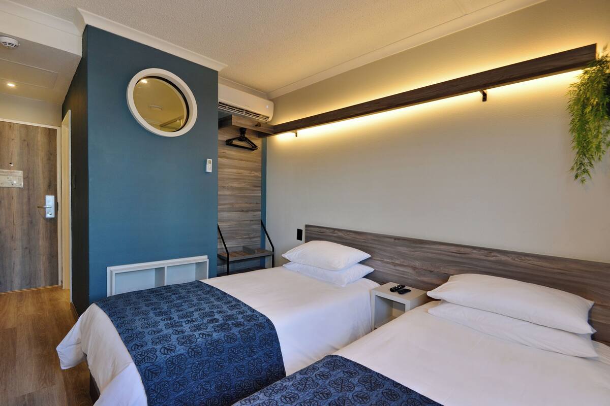 A hotel room with two beds, a round mirror, a wooden headboard, a nightstand, and a hanging plant.