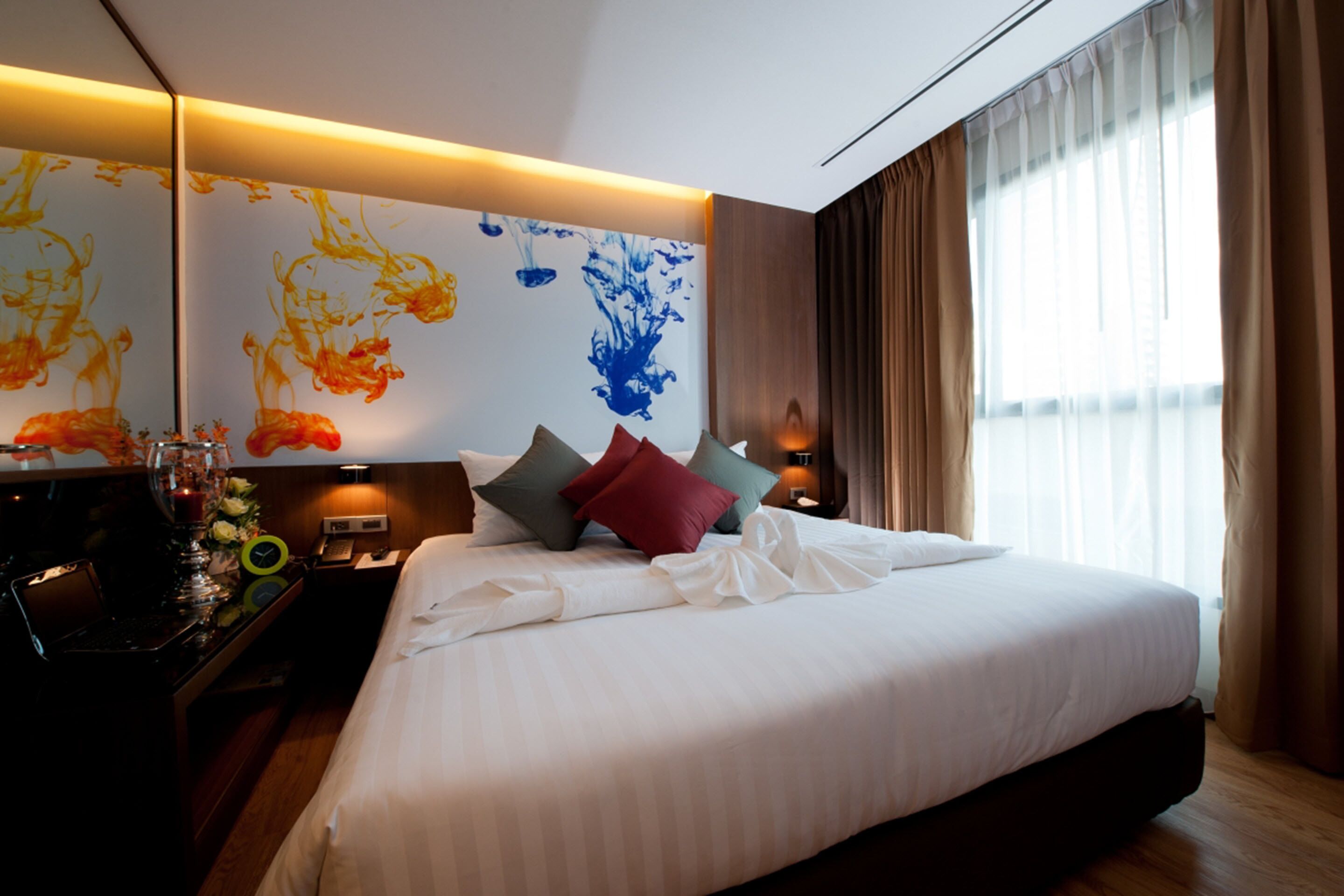 A modern hotel room with a large bed, a nightstand with a lamp, and a wall with abstract art.