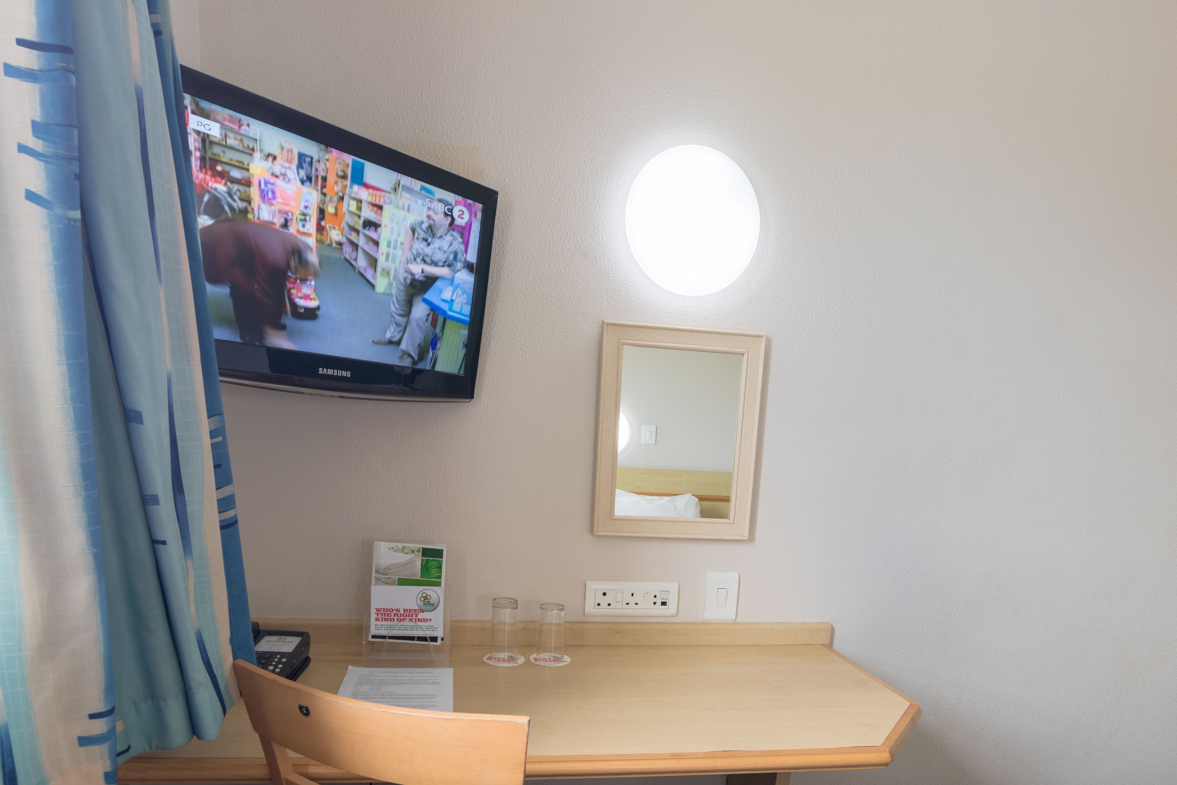 Habitación de hotel con una televisión montada en la pared, una mesa con una silla, un espejo y una mesita con un teléfono y documentos.