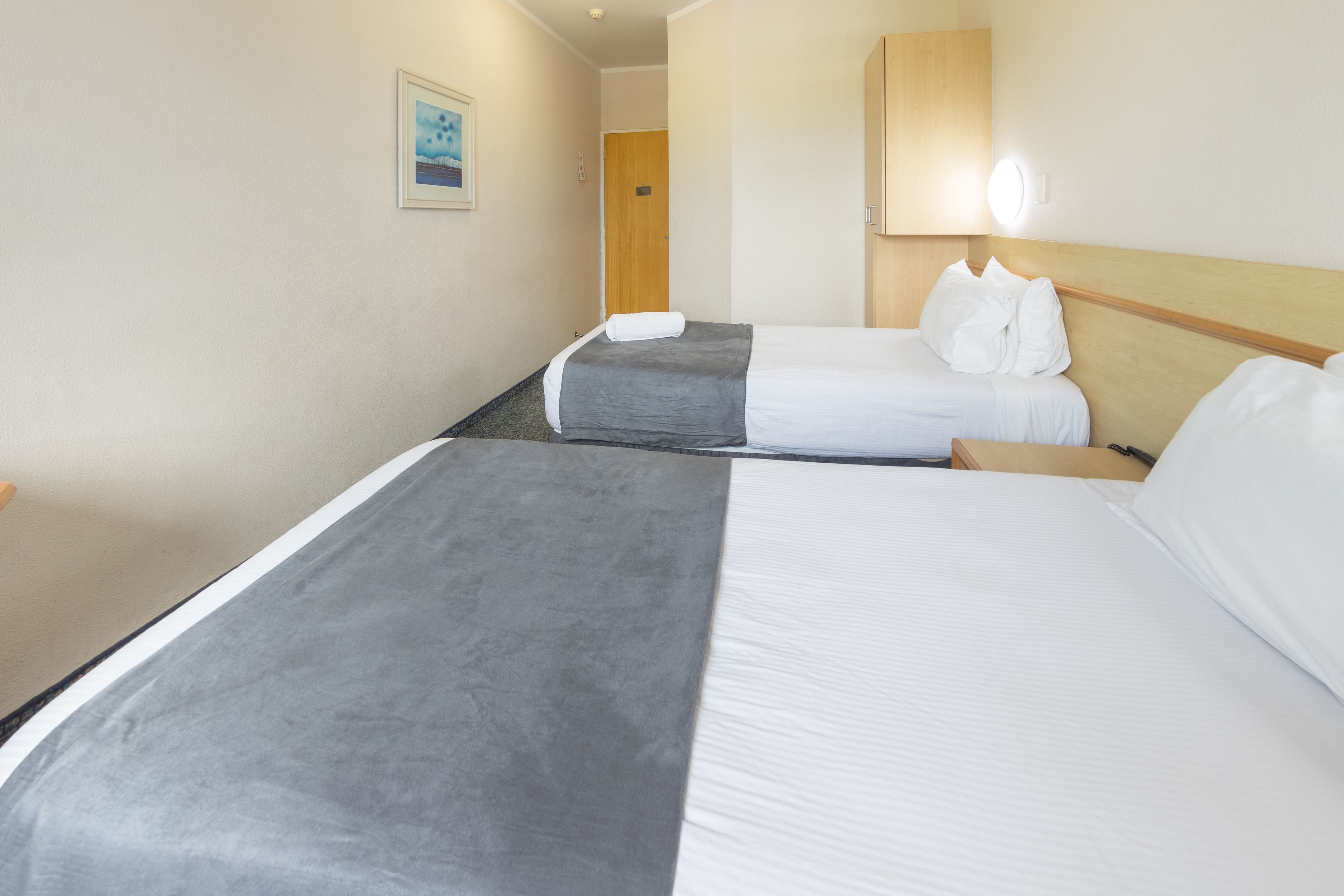 A hotel room with two beds, a wooden headboard, a painting on the wall, and a door in the background.