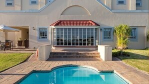 Uma área de piscina com um alpendre coberto, escadas que levam a um edifício e um gramado.