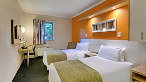 Quarto de hotel com duas camas, uma escrivaninha, uma cadeira, uma janela com cortinas, um espelho e uma estante com livros.