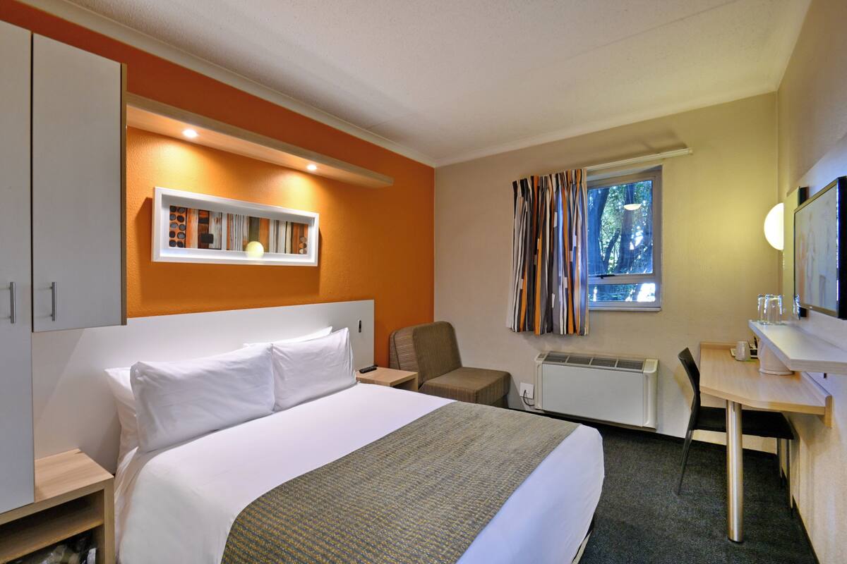 Habitación de hotel con cama, escritorio, silla, radiador, ventana con cortinas y estante con fotografías.