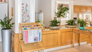 Un área de recepción con un escritorio de madera, plantas en macetas, un tablón de anuncios con un periódico y un cuadro grande en la pared.