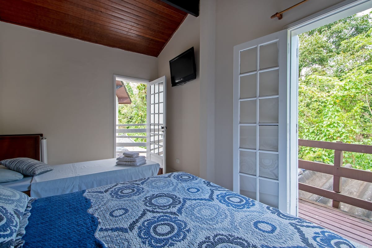 A bedroom with a bed, a television, a window with a view of greenery, and a balcony.