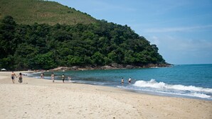 A beach with people walking along the shore, a sandy beach, and a forested hillside in the background. - Pauba Beach Hotel (São Sebastião)