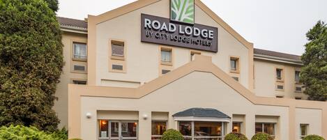 A building with a sign reading "Road Lodge by City Lodge Hotels." It features a modern design with a flat roof, multiple windows, and a landscaped entrance with small trees and shrubs.