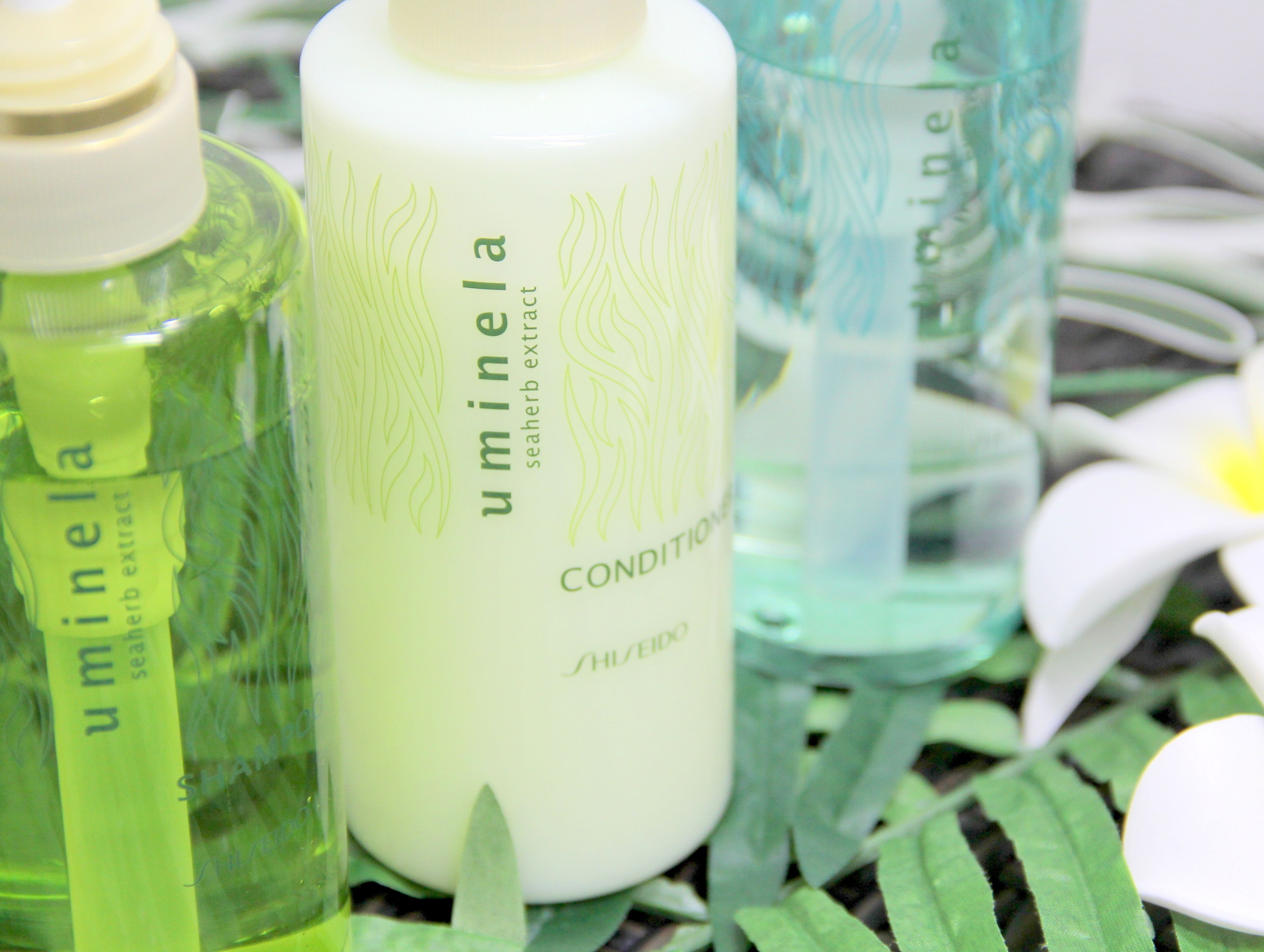 Eine Flasche 'Uminela'-Meerestrank-Konditionierer, umgeben von weiteren Flaschen, mit einer weißen Blume im Hintergrund.