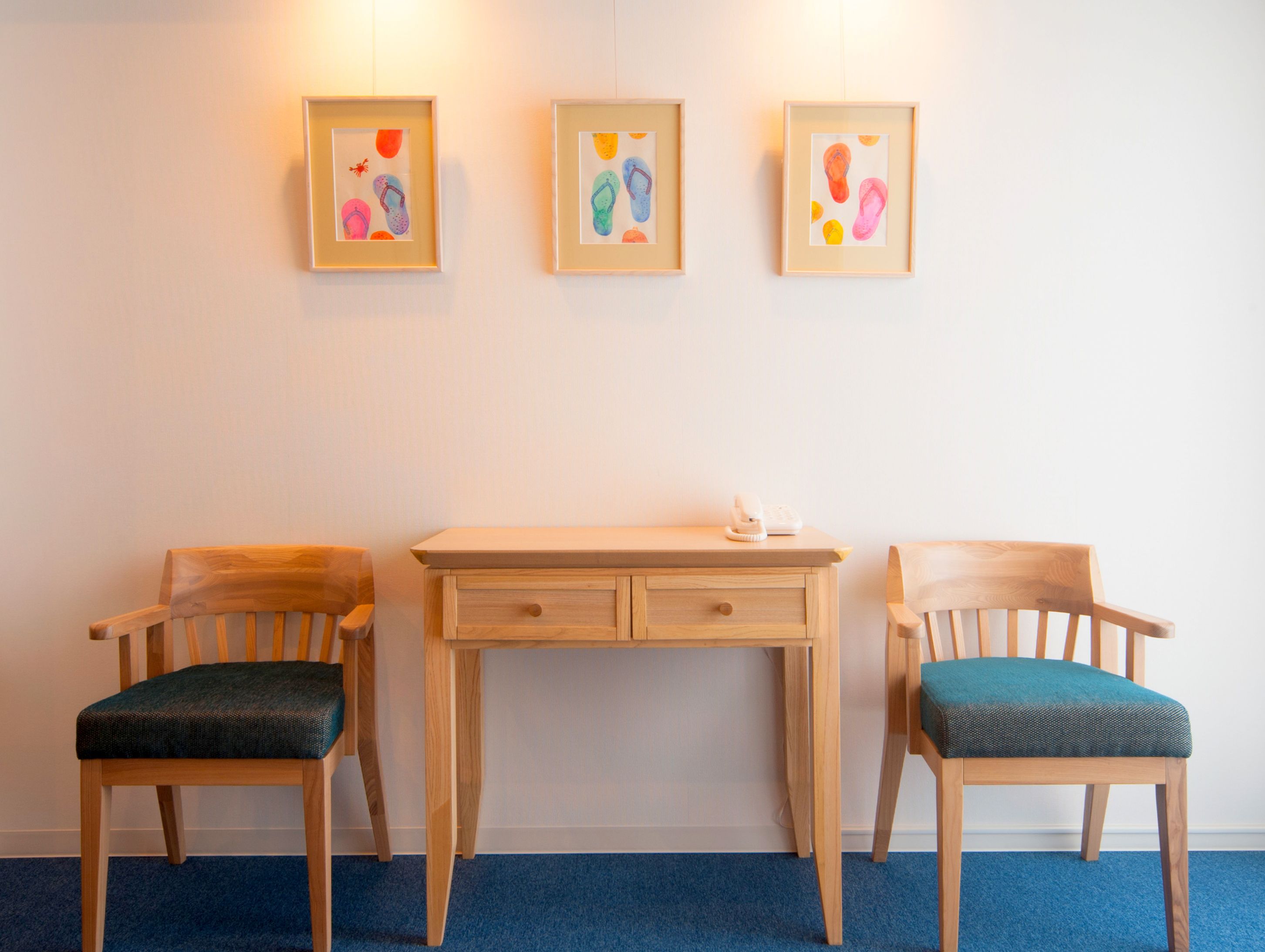 A room with two wooden chairs with teal cushions, a wooden desk, and three framed artworks on the wall.