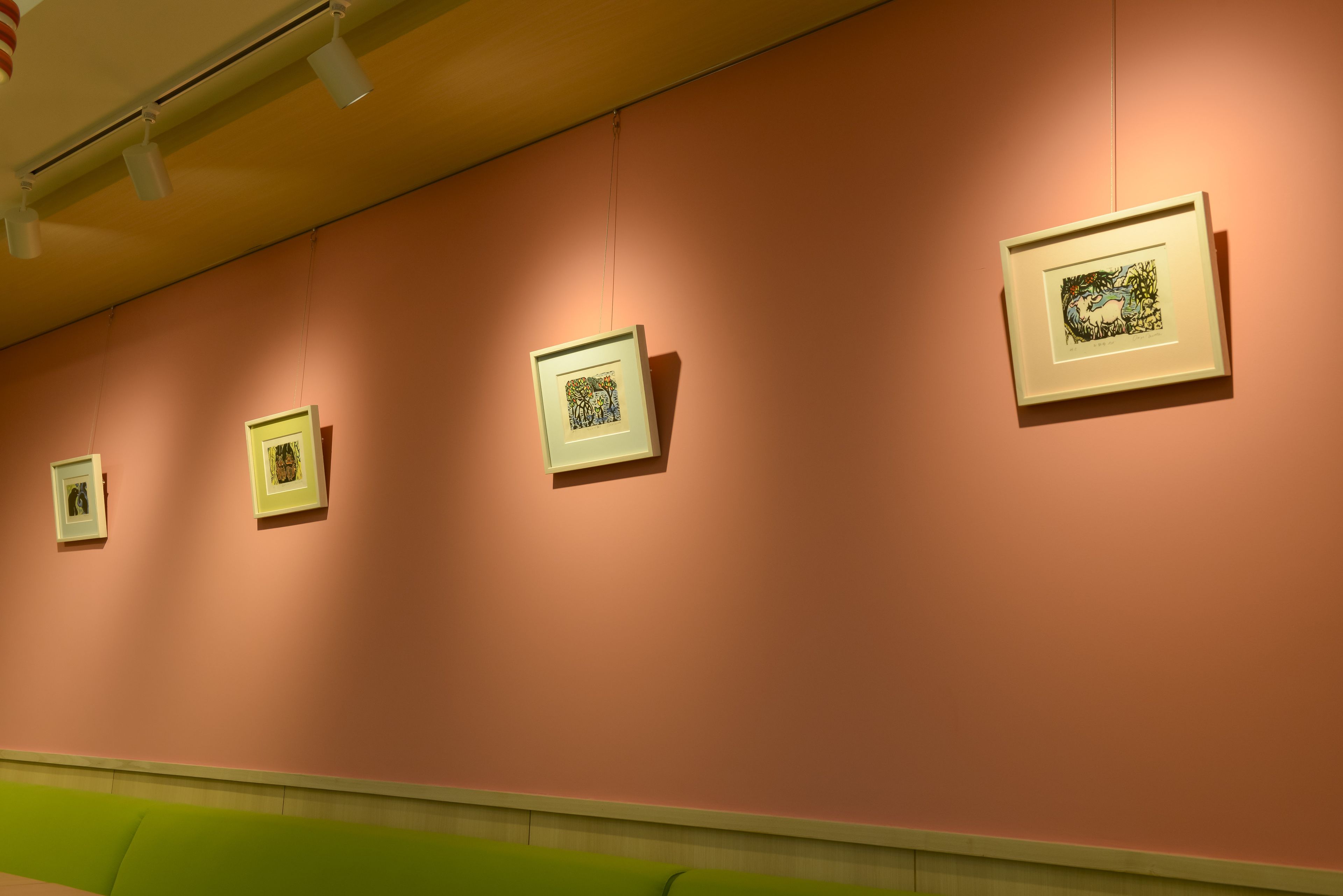 A gallery with framed artworks on a pink wall, a green bench, and a ceiling light fixture.