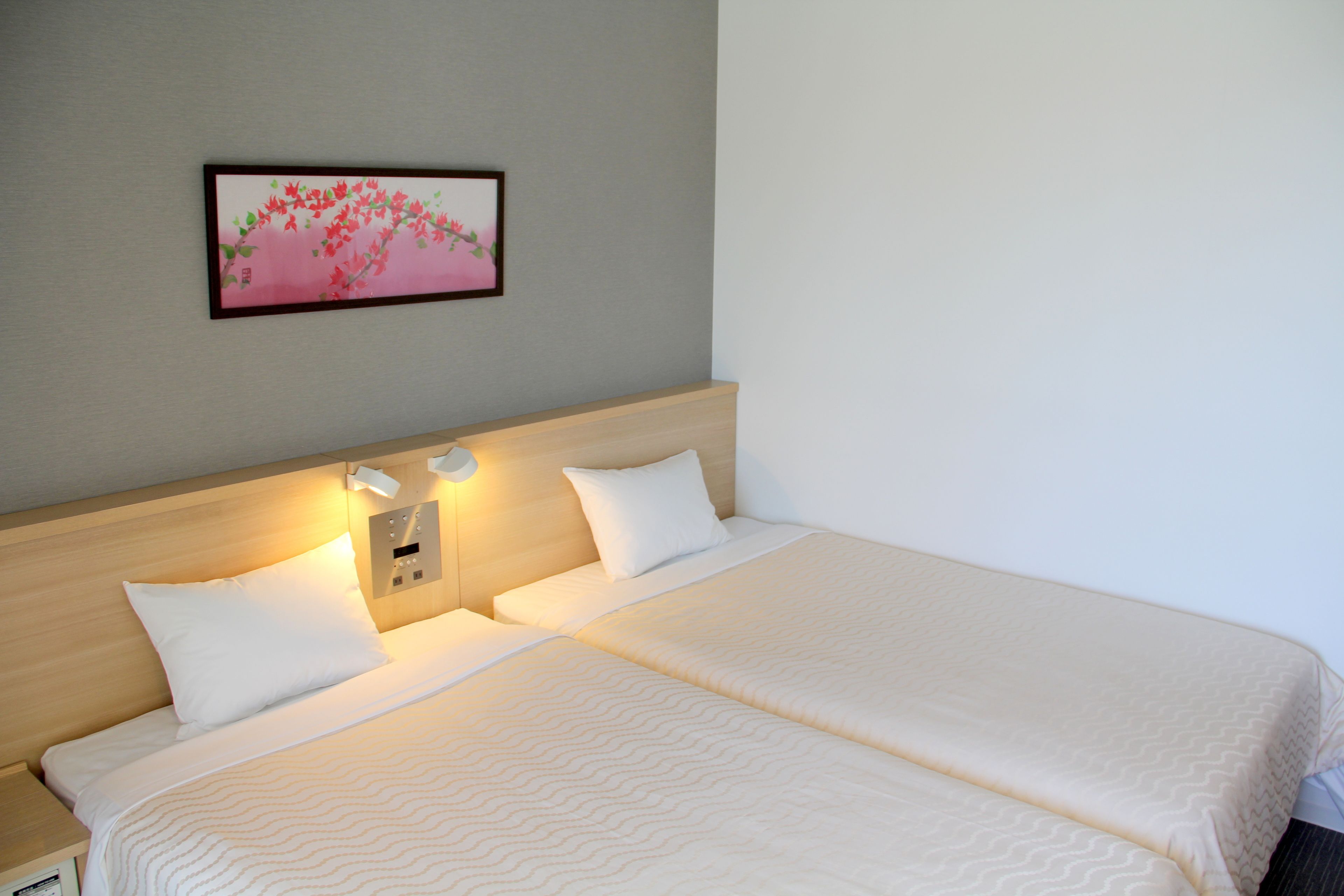 Habitación de hotel con dos camas, una mesita de noche, una lámpara de pared e imagen enmarcada.