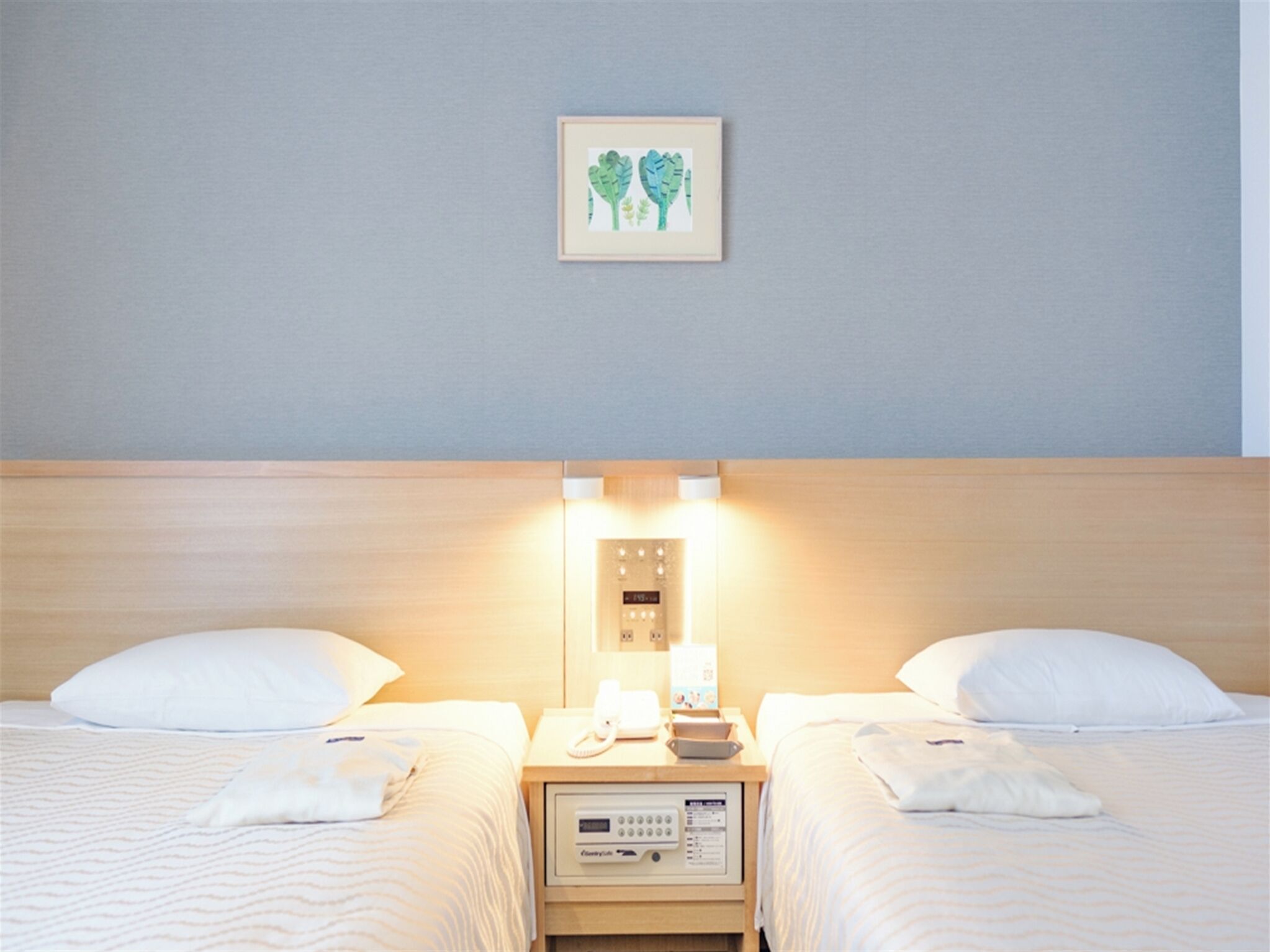 A hotel room with two beds, a bedside table with a phone and a lamp, and a wall-mounted control panel.