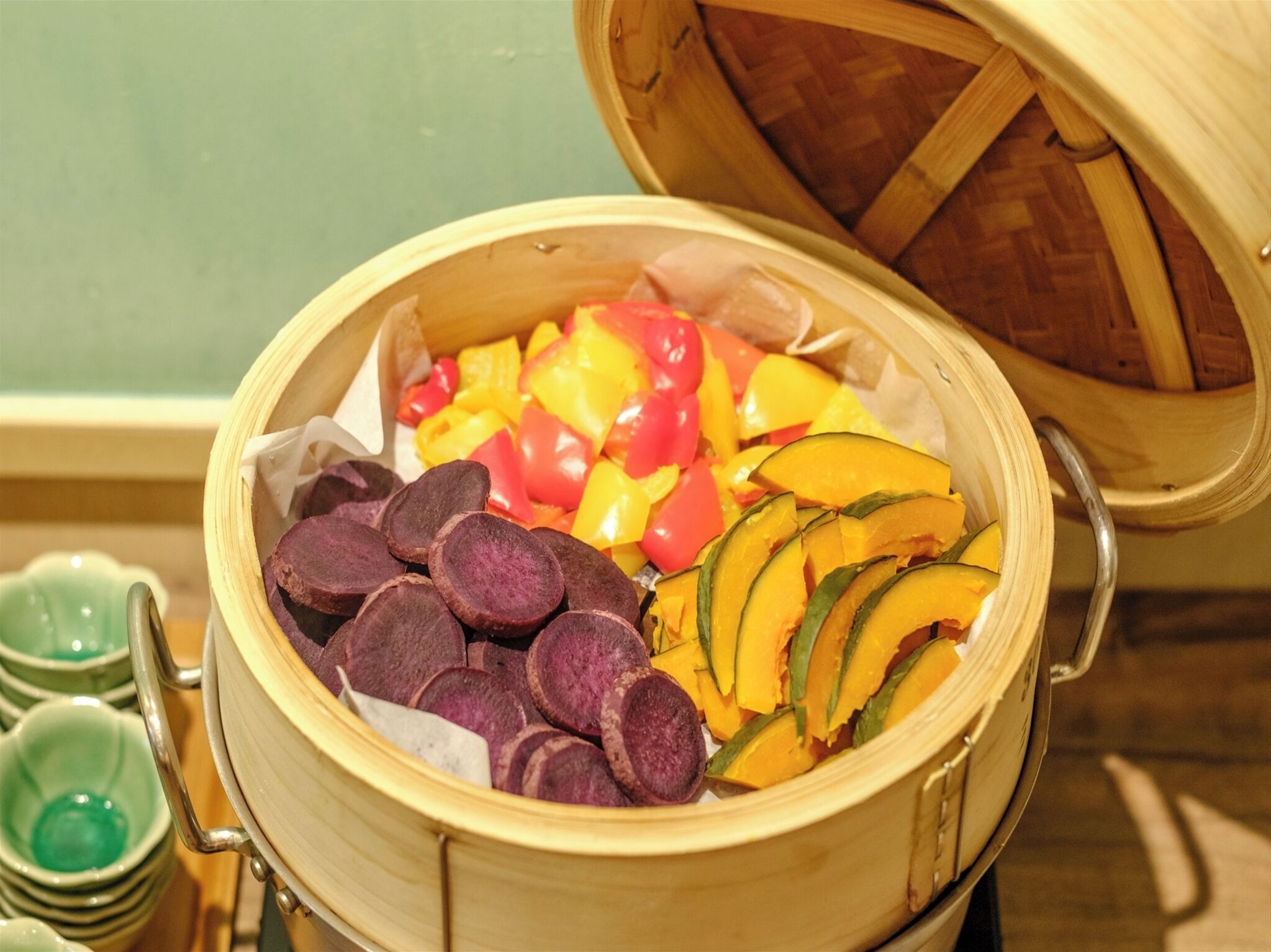 A bamboo steamer filled with sliced purple sweet potatoes, yellow and red bell peppers, and green zucchini.