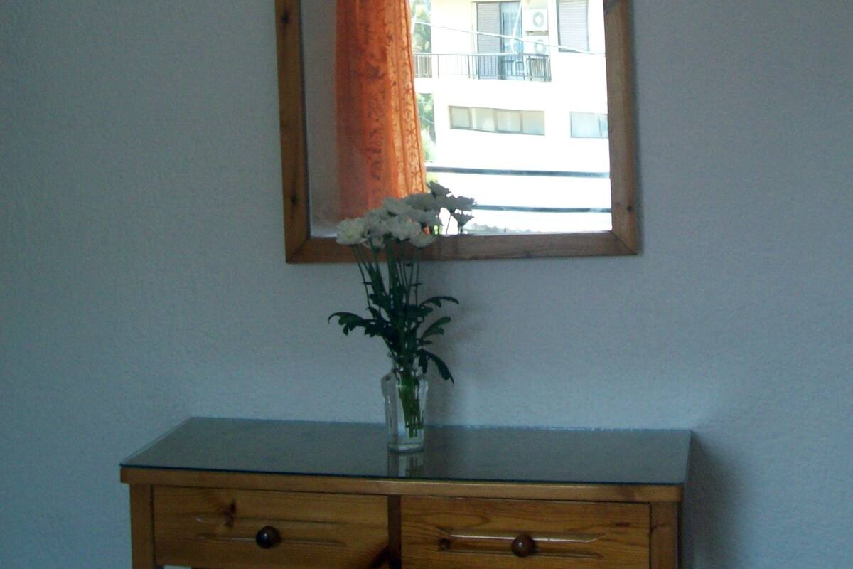 Ein Holztisch mit einem Spiegel darüber, eine Vase mit Blumen und ein Fenster mit Vorhang.