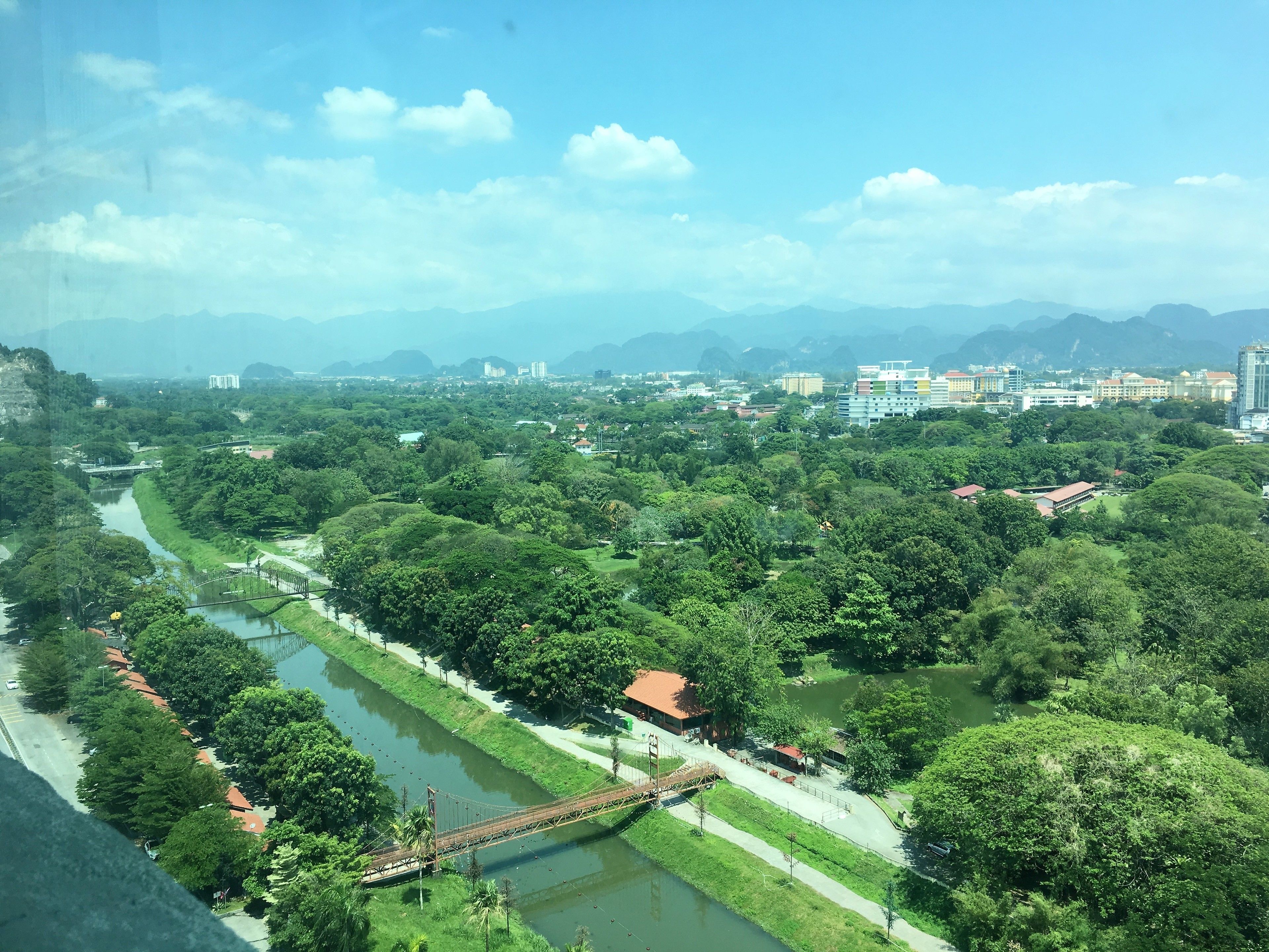 A river running through a park with a bridge, surrounded by greenery and buildings in the distance.