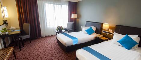 A hotel room with two beds, a desk, a chair, a lamp, and a window with curtains.