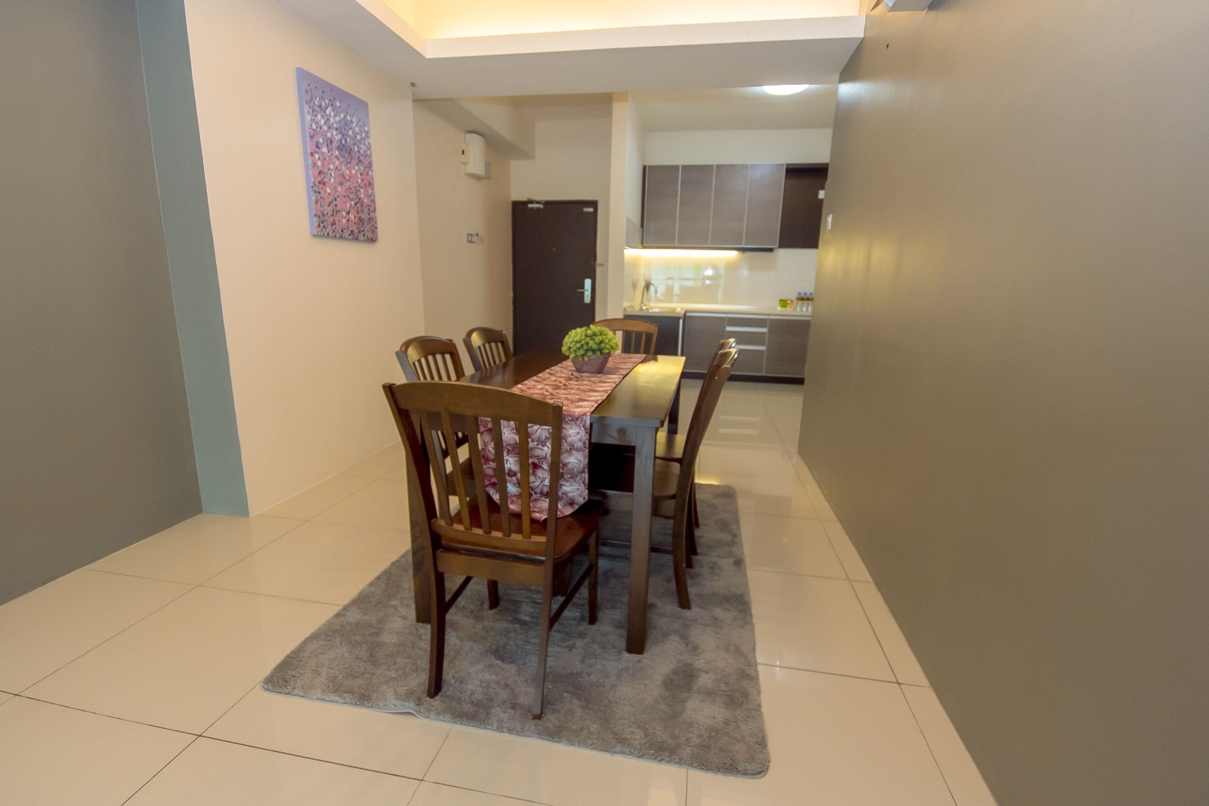 A dining area with a table, chairs, and a rug. There is a kitchen in the background with cabinets and appliances.