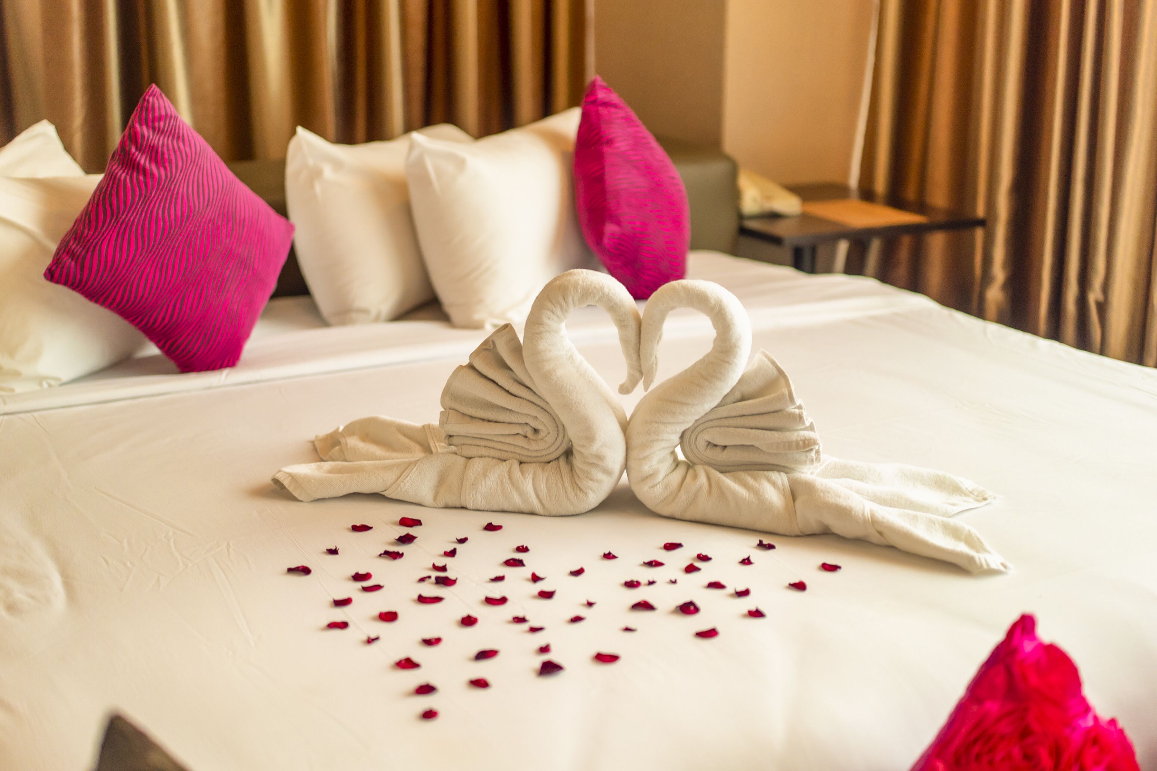A neatly made bed with a swan towel design and scattered rose petals.