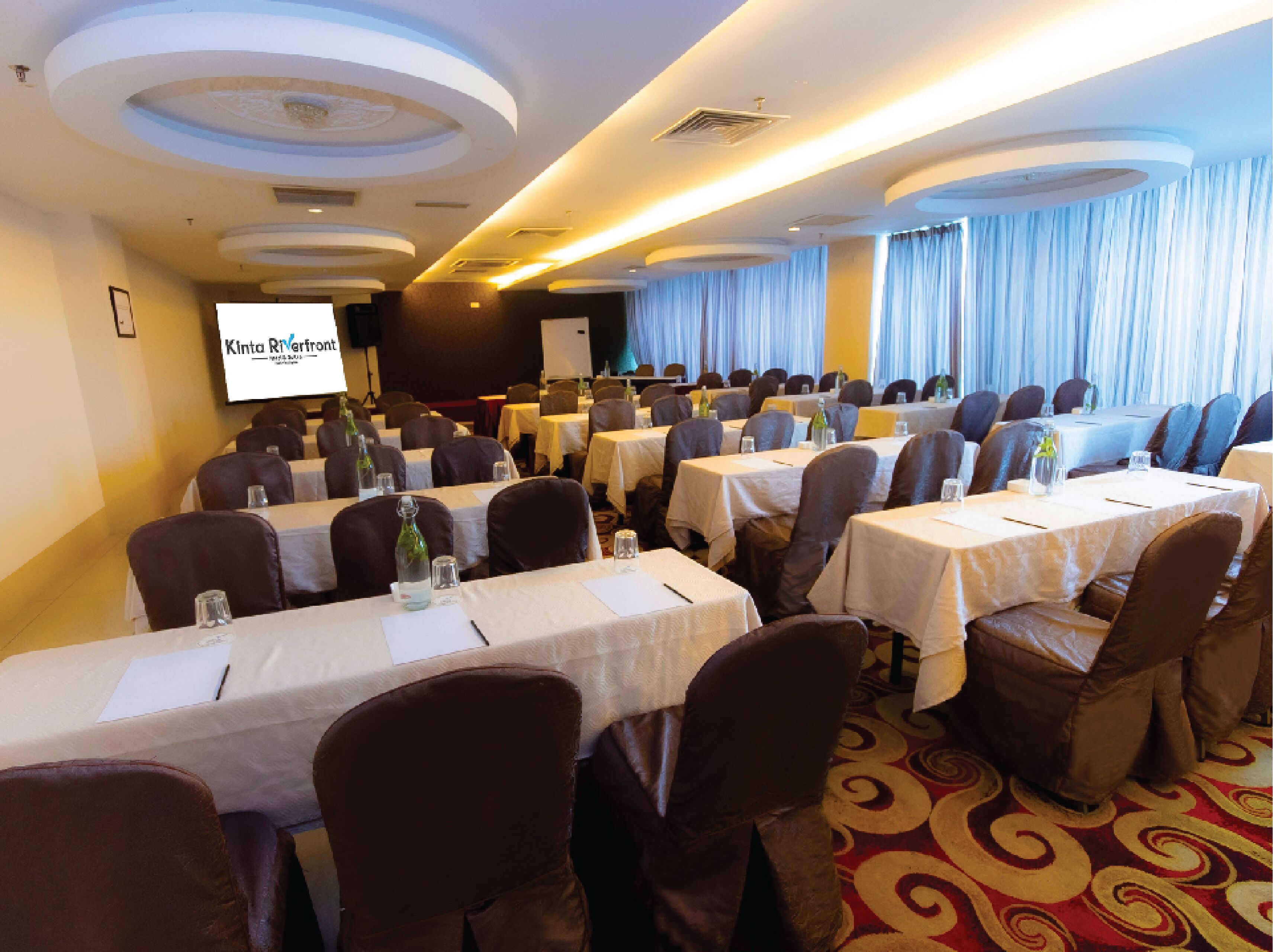 A conference room with tables set for a meeting, a projector screen, and a large window with curtains.