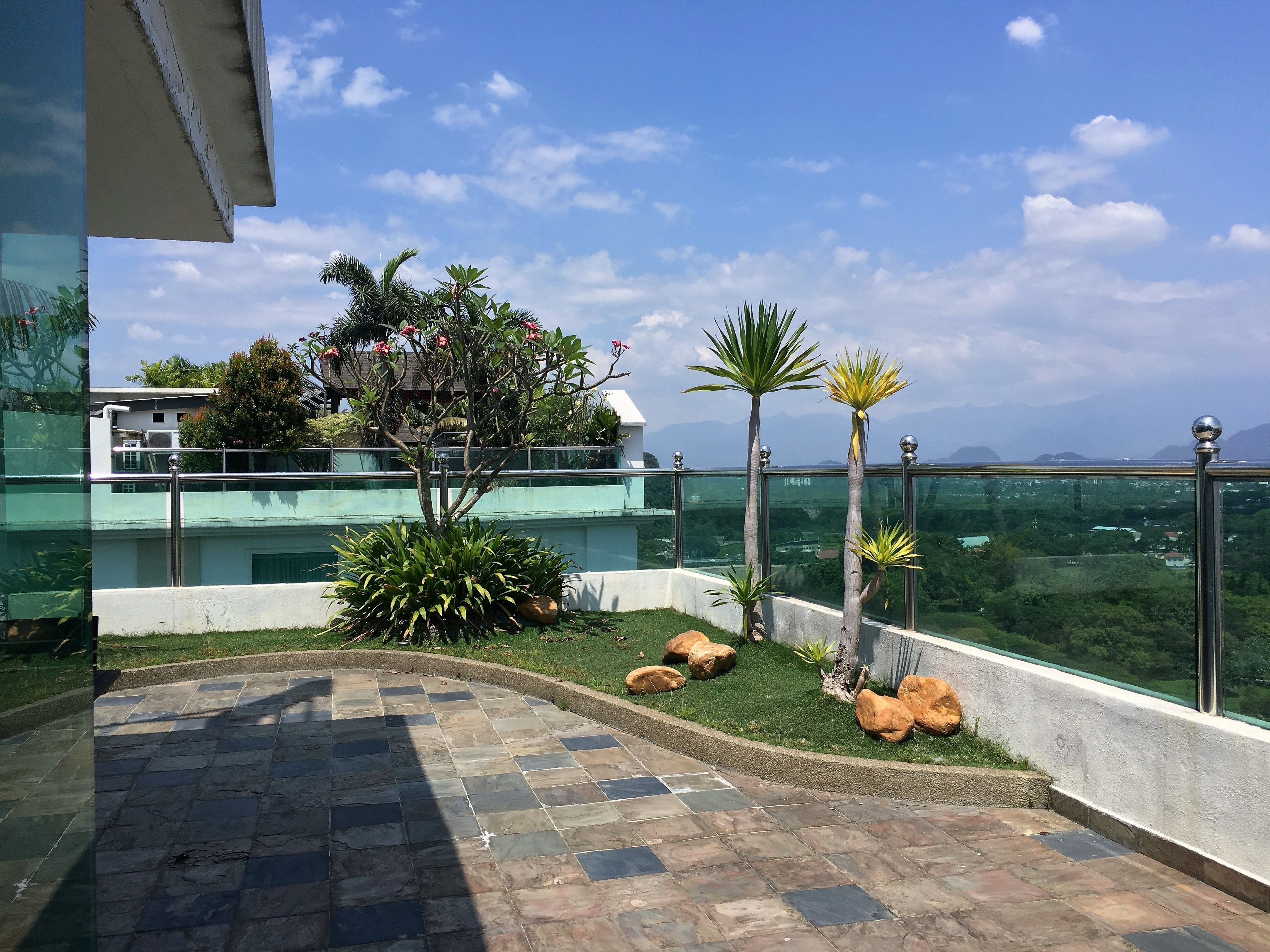 A rooftop terrace with a stone-tiled floor, a small garden with plants and rocks, and a view of a cityscape and mountains in the distance.